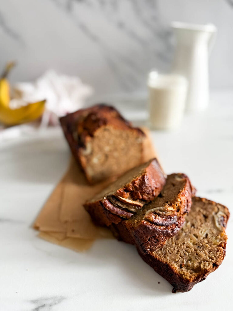 Banana Bread dorato e umido con fette disposte su carta da forno. Un bicchiere di latte e una caraffa bianca sfocati sullo sfondo. Un tocco rustico con una banana matura accanto.
