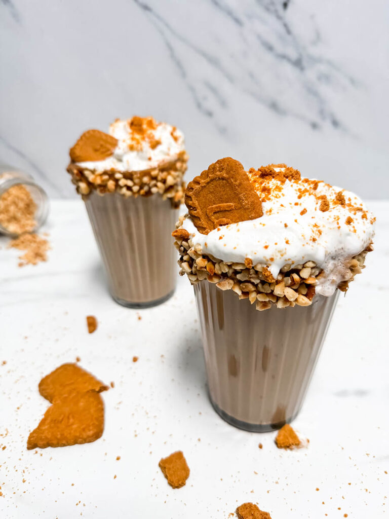 Due bicchieri di Biscoff Milkshake cremoso decorati con panna montata, biscotti Biscoff e granella di nocciole. Biscotti sbriciolati sparsi su un piano bianco marmorizzato. Un dolce indulgente e goloso.