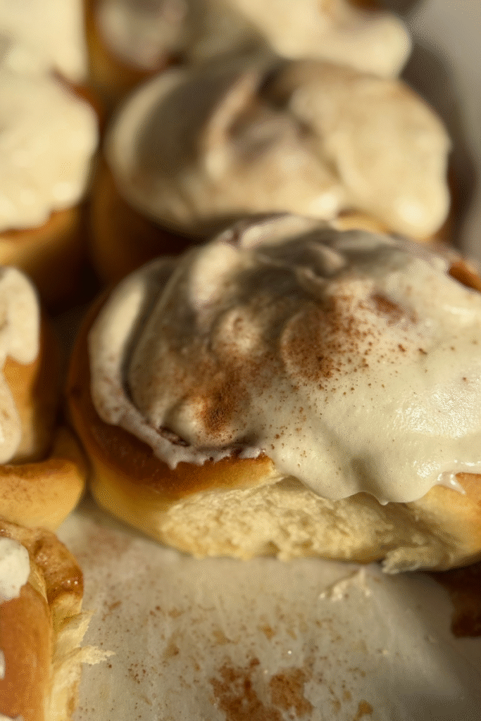 Primo piano di un cinnamon roll soffice ricoperto da una generosa glassa alla vaniglia, con una spolverata di cannella sopra. La luce calda evidenzia la consistenza morbida dell’impasto.