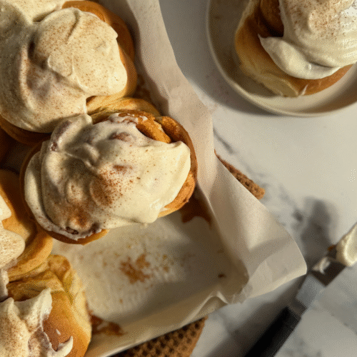 Vista dall’alto di una teglia di cinnamon roll glassati, con un roll servito a parte su un piattino. Il set è completato da un coltello e uno strofinaccio marrone su un piano in marmo.