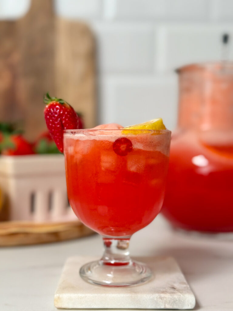 Primo piano di un bicchiere di strawberry lemonade con ghiaccio, guarnito con una fragola fresca e una fetta di limone. Sullo sfondo sfocato si intravede una caraffa con altra bevanda.
