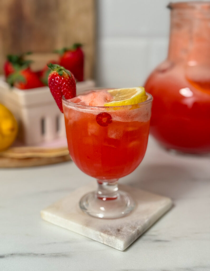 Bicchiere di strawberry lemonade servito su una base di marmo, decorato con una fragola intera e una fetta di limone. Sullo sfondo si notano limoni freschi e un cestino di fragole.